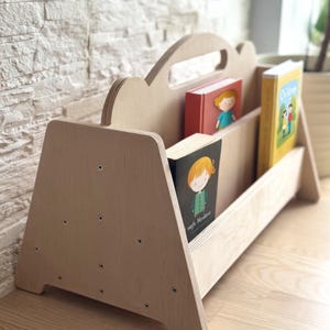Könnte beinhalten: Ein helles Kinderbücherregal aus Holz mit einer wolkenförmigen Oberseite. Das Bücherregal enthält drei farbenfrohe Kinderbücher. Das Bücherregal ist aus hellem Holz gefertigt und hat ein schlichtes, modernes Design.
