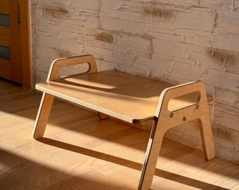 May include: A light wood bench with two handles on the top and a simple design. The bench is sitting on a light wood floor.