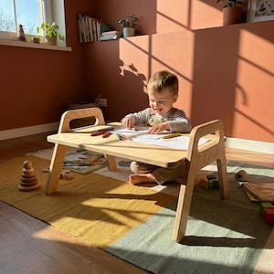 May include: A small, light-coloured wooden table with a handle on each end. A child is seated at the table, drawing with crayons on a piece of paper. The table is on a colourful rug. A wooden stacking toy is on the floor.