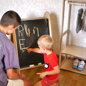 May include: Two children, one sitting and one kneeling, are using a chalkboard easel. The chalkboard is black and has white chalk writing on it. The writing says "RWM E". The child kneeling is pointing at the chalkboard with their right hand. The child sitting is looking at the chalkboard. There is a wooden clothes rack in the background.
