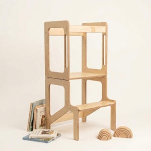 May include: A light-coloured wooden learning tower with a two-tier design, featuring a platform and a lower step. The structure is made of smooth plywood with open side panels. Several children's books and two wooden rainbow arches are nearby.