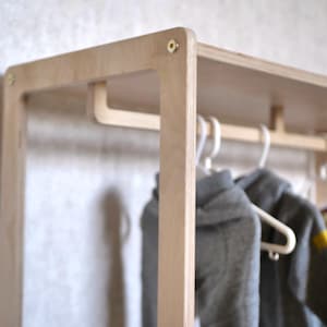 Könnte beinhalten: Ein hellfarbiger Kleiderständer aus Holz mit rechteckigem Rahmen. Der Ständer hat ein oberes Regal und eine untere Stange zum Aufhängen von Kleidung. Zwei graue Kapuzenpullover hängen an weißen Kleiderbügeln. Der Ständer ist aus hellfarbigem Holz gefertigt und hat messingfarbene Beschläge.