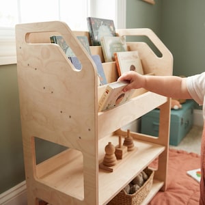 Puede incluir: Una estantería de madera clara con una sección superior para exhibir libros y un estante inferior que contiene un juguete apilable de madera y una cesta tejida. La unidad tiene asas recortadas en los paneles laterales y acabado natural.
