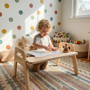 Könnte beinhalten: Ein Kindertisch aus hellem Holz mit integriertem Griff. Ein Kind zeichnet mit Buntstiften auf dem Tisch. Ein Holzspielzeug zum Stapeln und Buntstifte liegen auf dem Tisch. Der Raum hat eine farbenfrohe Polka-Dot-Wand.