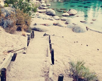 May include: A wooden staircase leads down to a sandy beach with clear blue water. The staircase is made of weathered wood planks and has a rustic look. The beach is surrounded by rocks and trees.