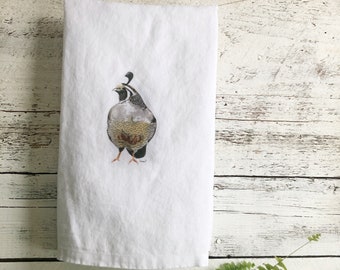 May include: White hand towel featuring a detailed watercolor illustration of a quail. The bird is depicted with gray, brown, and black feathers, standing on a white background. The towel is hanging on a thin string against a rustic, white-painted wooden backdrop.