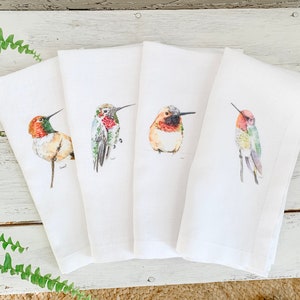 May include: Set of four white linen tea towels featuring watercolour hummingbird illustrations. Each towel depicts a different hummingbird species in various colours, including red, orange, green, and brown.
