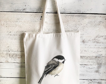 May include: A white canvas tote bag with a black and white watercolor illustration of a chickadee bird. The bird is facing left and has a black cap and bib.