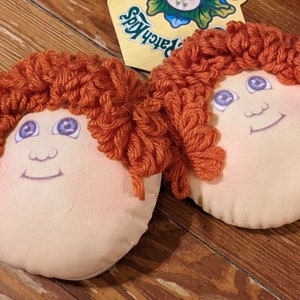 May include: Two plush dolls with red yarn hair and painted faces. The dolls have blue eyes and are smiling. The dolls are sitting on a wooden surface.