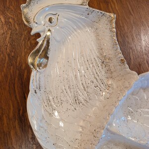May include: White ceramic rooster-shaped serving dish with gold accents. The dish features a detailed design of a rooster's head and body, with gold trim on the comb, beak, and edges. The surface has a speckled pattern.