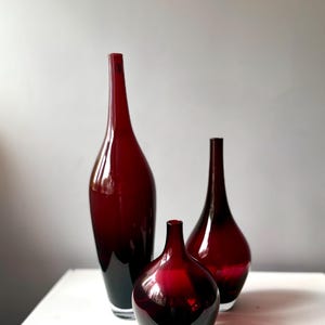 May include: Three decorative glass vases in varying sizes. The vases are a deep red colour, with a glossy finish. The tallest vase has a slender, elongated shape, while the other two have more rounded forms. The vases are displayed on a white surface.