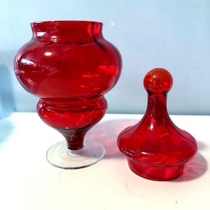 May include: A bright red glass apothecary jar and lid. The jar has a rounded body and a clear glass base. The lid has a spherical top. The objects are displayed on a white surface.