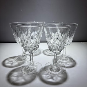 May include: Five clear crystal wine glasses with diamond-cut patterns. The glasses have a stemmed base and a starburst design on the foot. The glasses are arranged on a white surface with shadows.