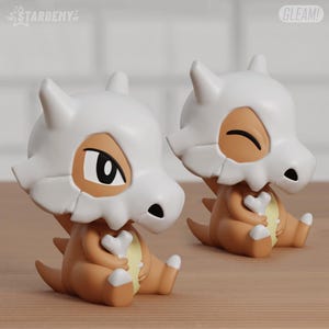 Könnte beinhalten: Zwei Cubone-Figuren, ein Pokémon-Charakter, werden gezeigt. Die Figuren sind hauptsächlich hellbraun und weiß, mit einem knochenförmigen Helm und einem Knochen in ihren Händen. Die Figuren sitzen. Die Texte "STARDENY" und "GLEAM!" sind sichtbar.