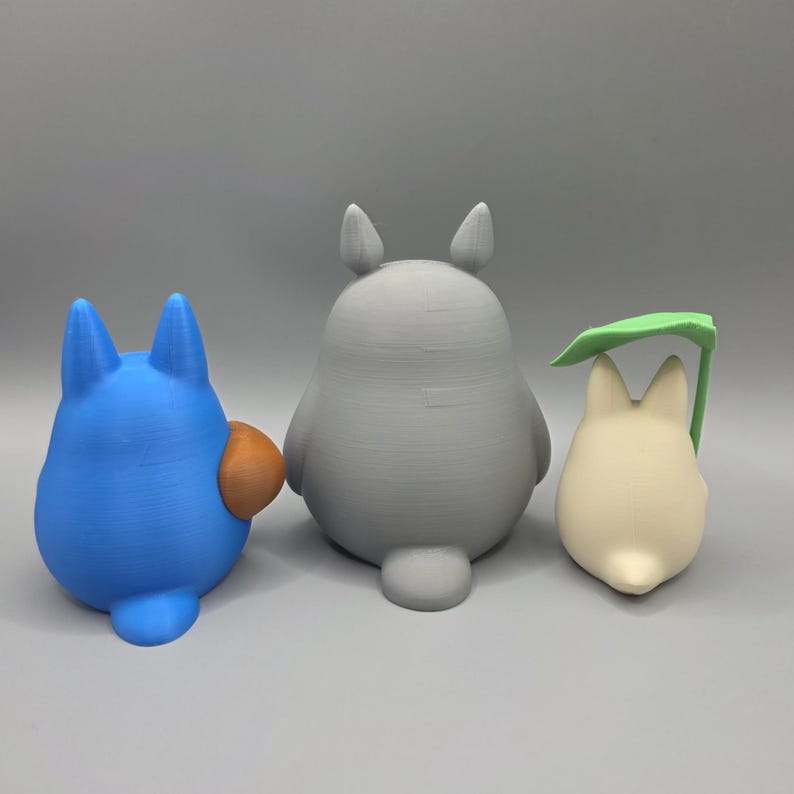 May include: Three 3D-printed figures of cartoon characters. The figures are a blue creature with an orange belly, a grey creature, and a cream-coloured creature holding a green leaf. The figures are on a grey surface.