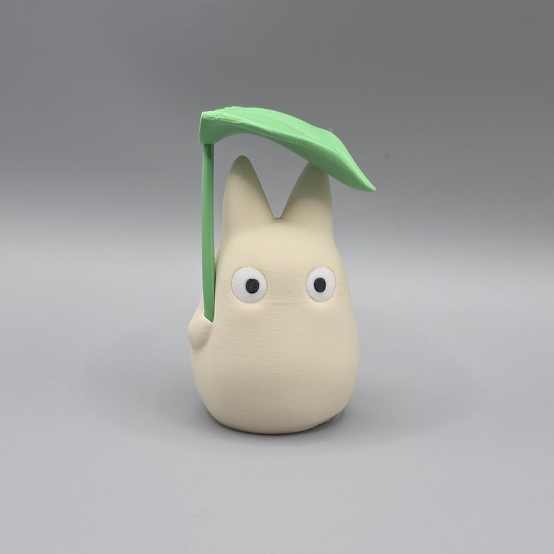 May include: A cream-coloured figurine of a cartoon character holding a green leaf overhead. The character has large black eyes and pointed ears. The leaf is supported by a green stem, creating a whimsical umbrella effect.
