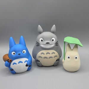 May include: Three figurines from the animated film, My Neighbour Totoro, are displayed. A blue Totoro holds a small brown sack. A grey Totoro smiles, and a small white creature holds a green leaf overhead. The figures are on a grey background.
