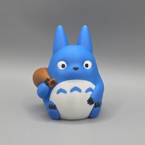 May include: A blue figurine resembling Totoro, holding a small brown sack. The figure has large white eyes, a white belly with three curved markings, and pointed ears. The figurine is set against a plain grey backdrop.
