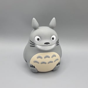 May include: A grey figurine of Totoro, a character from the animated film My Neighbour Totoro. The figure has a large, smiling face with black whiskers and a cream-coloured belly. It has pointed ears and is standing upright.