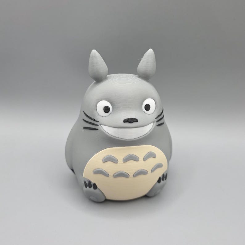 May include: A grey figurine of Totoro, a character from the animated film My Neighbour Totoro. The figure has a large, smiling face with black whiskers and a cream-coloured belly. It has pointed ears and is standing upright.