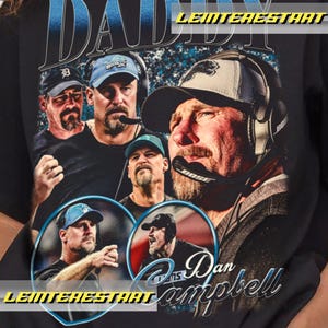 May include: Black t-shirt featuring a graphic of Dan Campbell, with the word "DADDY" in blue and white. The design includes multiple images of Campbell, with the name "Dan Campbell" in a cursive font.