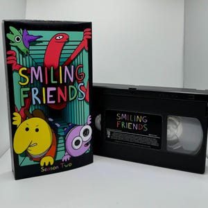 May include: A black VHS tape with a colorful cartoon image of a red worm-like character, a yellow character with a cowboy hat, and a purple character with large eyes. The tape is labeled "Smiling Friends" and the box is labeled "Smiling Friends Season Two".