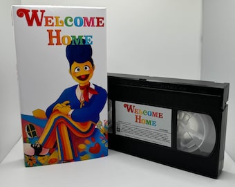 May include: A VHS tape with a colorful cover featuring a cartoon character wearing a blue suit and rainbow pants. The cover reads "Welcome Home".