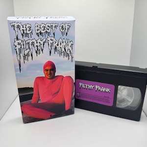May include: A VHS tape and box set with the text "THE BEST OF FILTHY FRANK." The box art depicts a person in a pink bodysuit and beanie. The VHS tape has a purple label with the text "FILTHY FRANK."