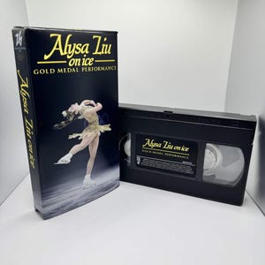 May include: A black VHS tape and box set featuring the text "Alysa Liu on ice - Gold Medal Performance." The box shows a figure skater in a gold costume. The tape and box are on a white surface.