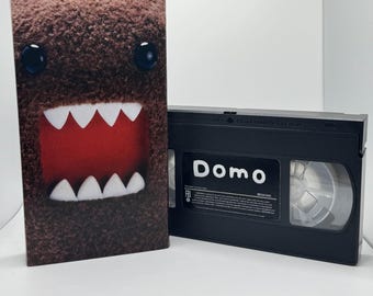 May include: A VHS tape with a brown Domo character on the cover. The tape is labeled "Domo" and has a black case.