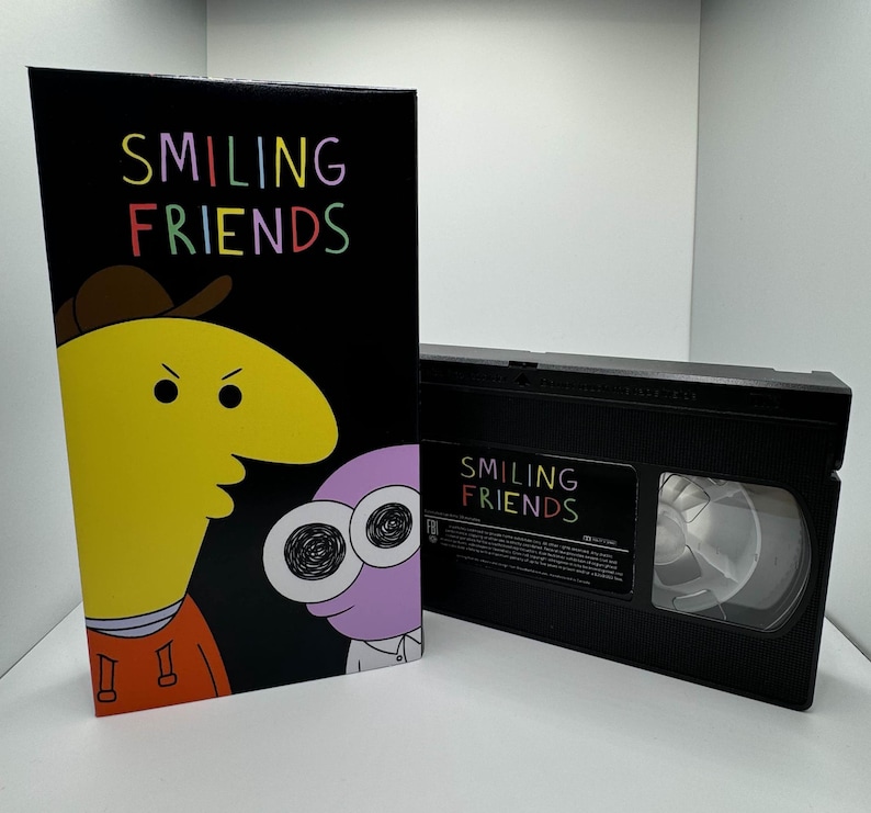 May include: A VHS tape with a black case and a label that says "Smiling Friends". The label has a rainbow color scheme. The cover art features two cartoon characters, one yellow with a black hat and the other purple with large eyes.