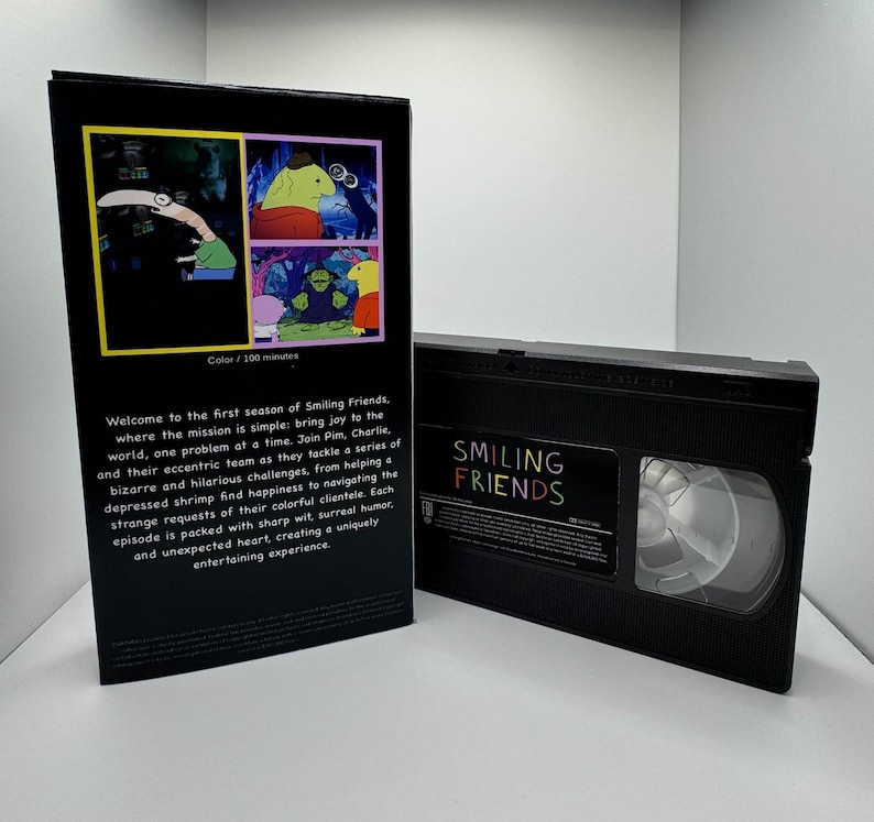 May include: A VHS tape case with a black background and a colorful image of cartoon characters. The case has the title "Smiling Friends" and the text "Color/100 minutes".