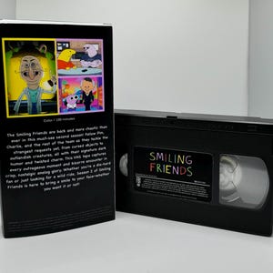 May include: A VHS tape case for the animated series "Smiling Friends". The front of the case features a black background with the title "Smiling Friends" in rainbow letters. The back of the case features a black background with a description of the show and a series of colorful cartoon stills.