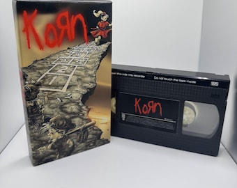 May include: A VHS tape case and cassette with the band Korn's name in red lettering. The case features a black and white illustration of a girl in a red dress walking on a cracked path with a hopscotch game drawn on it. The cassette has the band's name in red lettering.
