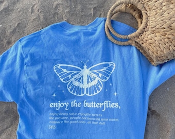 May include: A light blue t-shirt featuring a white graphic of a butterfly and the text "enjoy the butterflies, enjoy being naive, enjoythe nerves, the pressure, people not knowing your name, embrace the good ones, all that stuff" and "DR3".