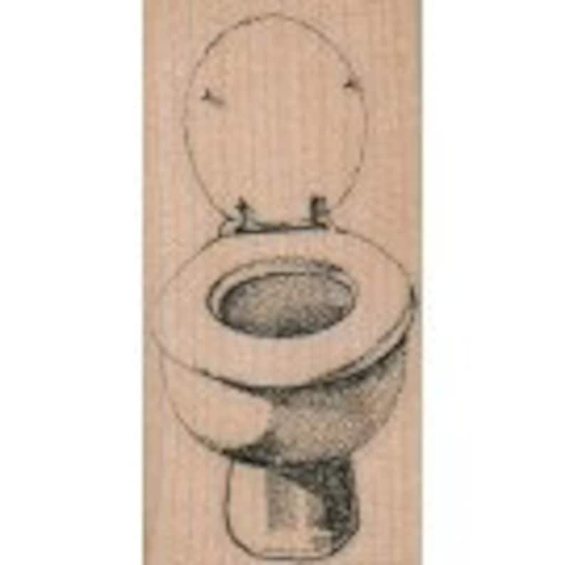 May include: A wooden rubber stamp with a black and white illustration of a toilet with the lid up.