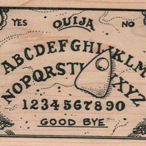 Puede incluir: Un tablero de Ouija de madera con letras negras y una planchette. El tablero presenta el alfabeto, los n&uacute;meros del 1 al 9 y el 0, y las palabras "S&iacute;", "No", "Adi&oacute;s" y "Ouija".