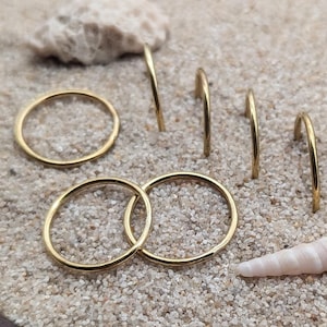 Op de afbeelding: Vijf gouden ringen in verschillende maten zijn gerangschikt op een houten dienblad gevuld met zand. De ringen zijn gerangschikt in een halve cirkelvorm, met de grootste ring bovenaan en de kleinste ring onderaan. Het dienblad is gemaakt van donker hout met een lichtbruine rand.