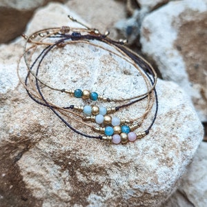 May include: Set of four bracelets with brown, black, and gold cords. Each bracelet features small, colourful beads in shades of blue, green, and pink.