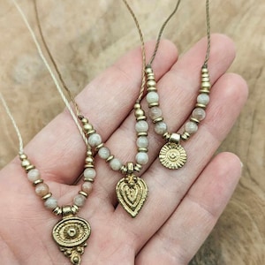 Peut inclure: Trois colliers avec des pendentifs dorés et des chaînes de perles. Les pendentifs ont des formes différentes, dont un ovale, une goutte et un motif de soleil. Les perles sont de couleur beige clair. Les colliers sont présentés sur une surface en bois.