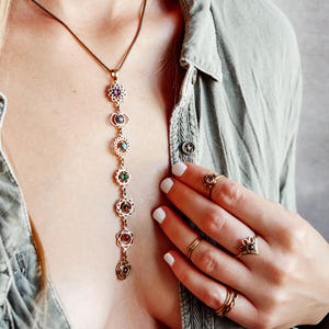 Op de afbeelding: Een gouden ketting met zeven chakra-symbolen en edelstenen. Elk symbool is een mandala met een edelsteen in een andere kleur in het midden. De ketting wordt gedragen door een persoon met een groen shirt.