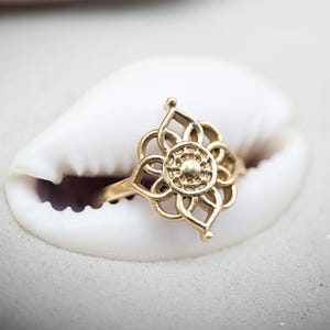 以下が含まれることがあります： 花柄のデザインが施されたゴールドカラーのリング。中央の円形要素と花びらのような形が特徴です。白い貝殻の上に置かれ、質感と色のコントラストを生み出しています。リングの複雑なディテールがクローズアップで強調されています。