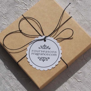 May include: A square, light brown gift box tied with black twine. A white, scalloped-edge tag is attached, bearing the text "Fourseasons Fragrance.com" in black. The box is presented on a textured, light-colored surface.