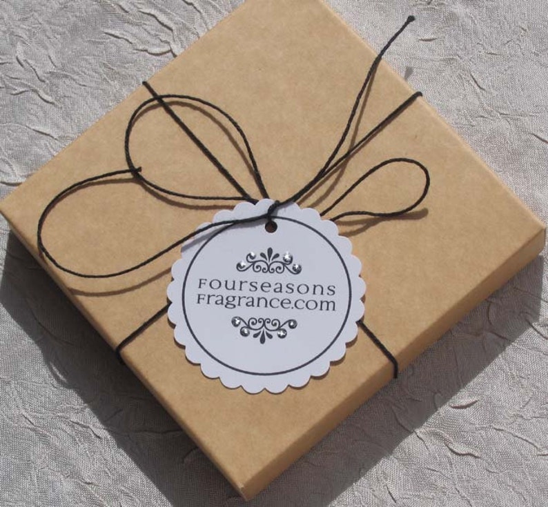 May include: A square, light brown gift box tied with black twine. A white, scalloped-edge tag is attached, bearing the text "Fourseasons Fragrance.com" in black. The box is presented on a textured, light-colored surface.