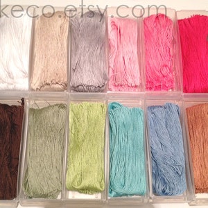 K&ouml;nnte beinhalten: Eine Sammlung von 16 Spulen mit Stickgarn in verschiedenen Farben, darunter Lila, Braun, Gr&uuml;n, Hellgr&uuml;n, Blau, Hellblau, Rosa, Rot, Schwarz, Wei&szlig;, Beige und Mehrfarbig. Die Spulen sind in zwei Reihen zu je acht in durchsichtigen Plastikbeh&auml;ltern angeordnet.