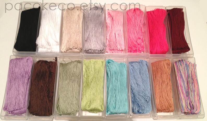 K&ouml;nnte beinhalten: Eine Sammlung von 16 Spulen mit Stickgarn in verschiedenen Farben, darunter Lila, Braun, Gr&uuml;n, Hellgr&uuml;n, Blau, Hellblau, Rosa, Rot, Schwarz, Wei&szlig;, Beige und Mehrfarbig. Die Spulen sind in zwei Reihen zu je acht in durchsichtigen Plastikbeh&auml;ltern angeordnet.