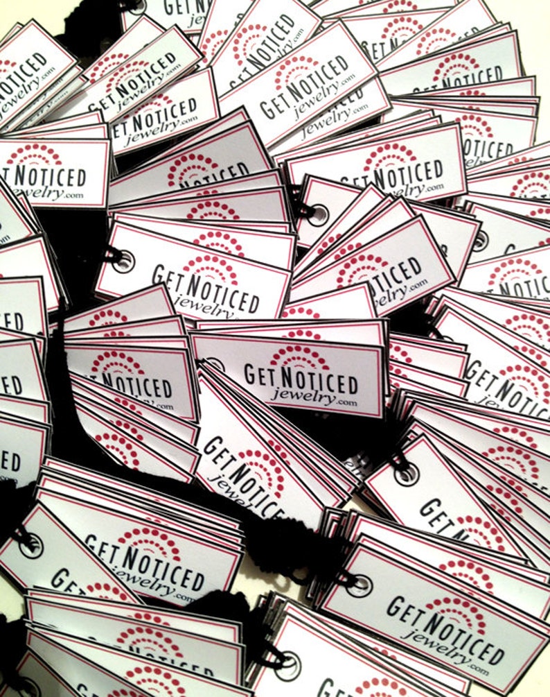 K&ouml;nnte beinhalten: Ein Stapel wei&szlig;er Geschenkanh&auml;nger mit schwarzen R&auml;ndern und roten Punkten. Die Anh&auml;nger haben den Text "Get Noticed Jewelry.com" aufgedruckt.