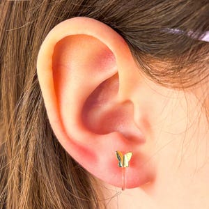 May include: A gold butterfly earring with a clear plastic post. The earring is worn in the left ear of a person with brown hair.