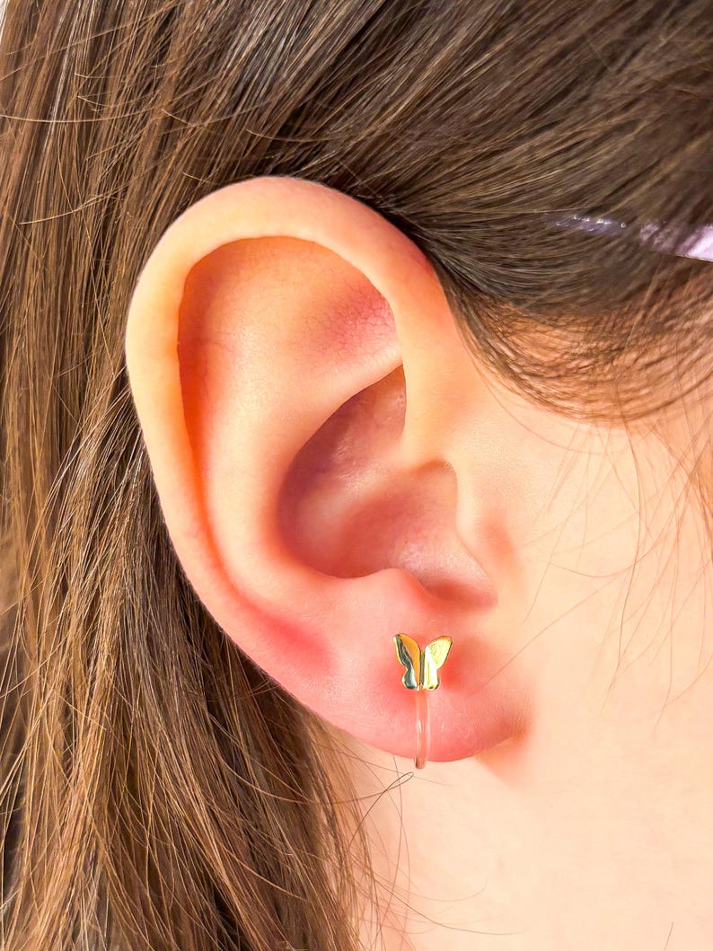 May include: A gold butterfly earring with a clear plastic post. The earring is worn in the left ear of a person with brown hair.