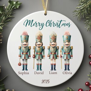 May include: A white Christmas ornament with a gold string, featuring four toy soldier figures. The ornament reads "Merry Christmas" in green script, with names and the year 2025 below the soldiers. The soldiers are in teal, red, and gold.